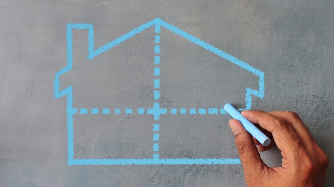 Una mano sta disegnando con un gessetto azzurro su una lavagna scura la sagoma di una casa divisa in quattro sezioni da linee tratteggiate, a simboleggiare la divisione ereditaria di un bene immobiliare. Leggi l'articolo per scoprire come facilitare la Divisione Ereditaria e prevenire i conflitti tra eredi.